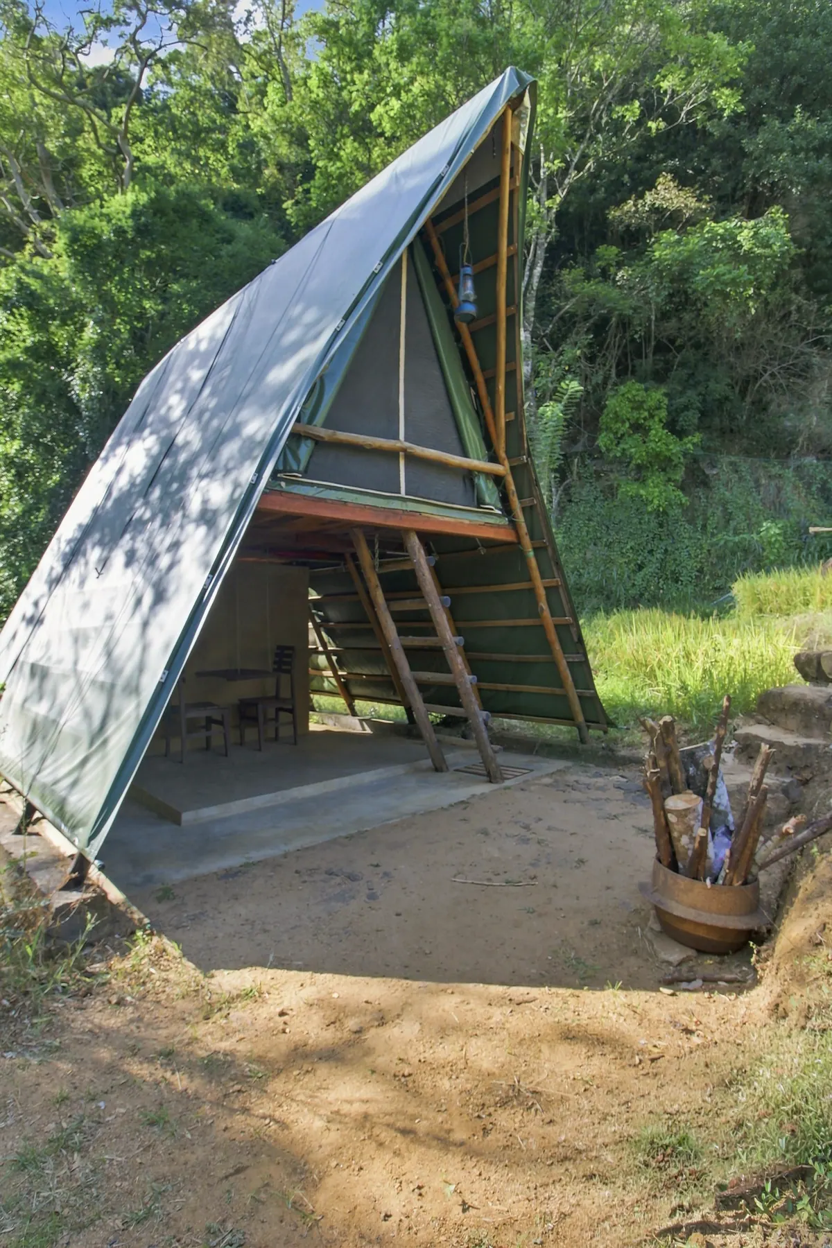 An A-frame glamping cabin in green canvas, its ladder up to the sleeping deck, against the forest