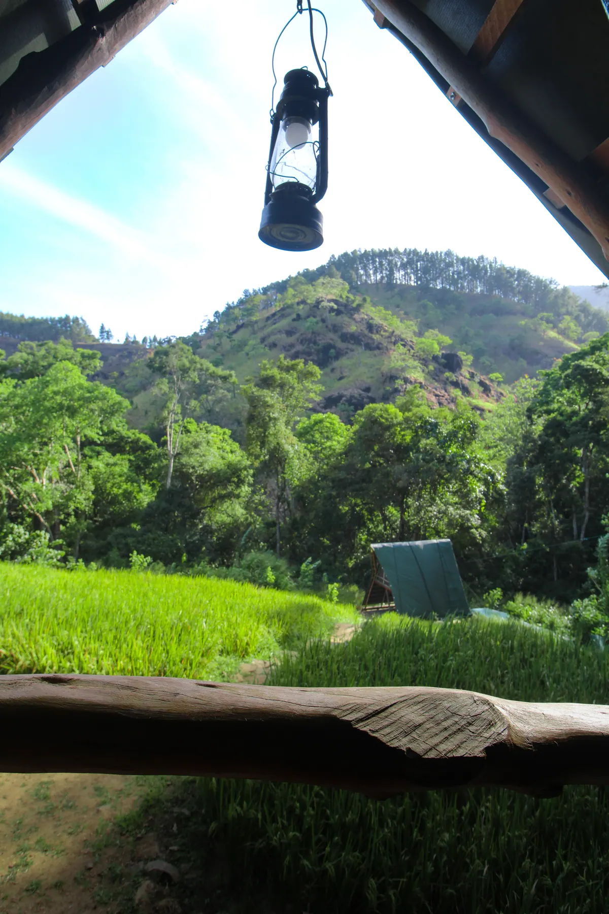 A hurricane lantern hanging in the open end of a cabin, the paddy and hills framed beyond